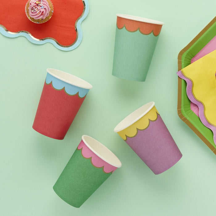 Colorful paper cups in red, green, purple, and mint green with white scalloped trim arranged on a pale green background.