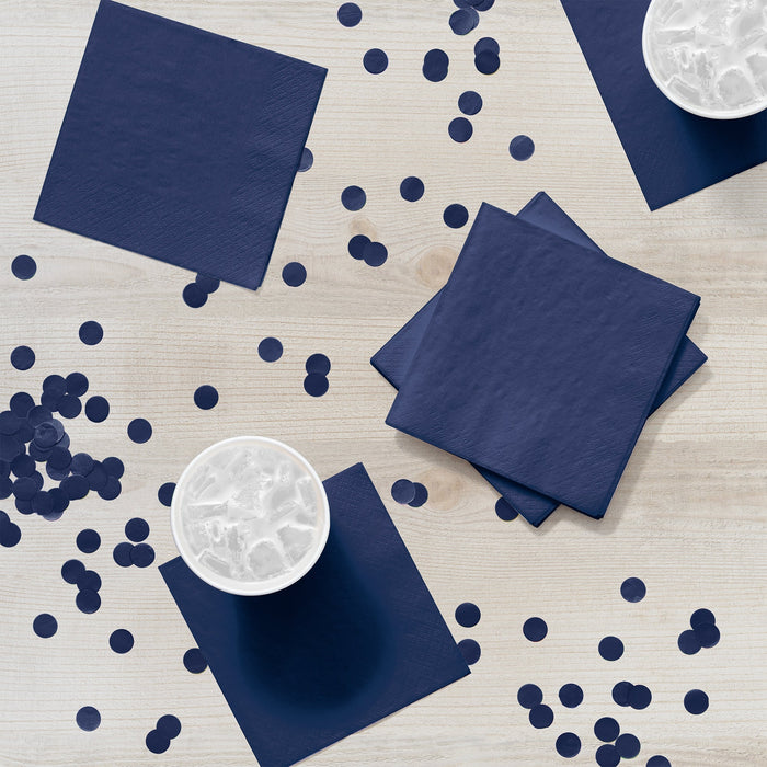 Navy blue paper napkins arranged on a light wood surface with scattered dark blue confetti dots, white cups with ice, and stacked napkins creating a party display.