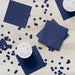 Navy blue paper napkins arranged on a light wood surface with scattered dark blue confetti dots, white cups with ice, and stacked napkins creating a party display.