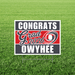 Red, white, and gray yard sign with "Congrats Grad 2026" text and "Owyhee" at bottom, displayed on grass with metal stake supports.