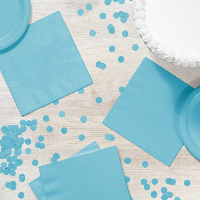 Pastel blue luncheon napkins arranged on a white wood surface with matching blue plates, confetti dots, and decorative white fringe trim visible in corners.