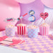 Four frosted cupcakes in striped and checkered wrappers in pink, purple, and blue sit on a purple and pink checkered mat with themed toppers including a "Pamper Party" eye mask, makeup brush, compact mirror, and heart-shaped pick.