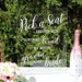 Clear acrylic wedding seating sign with white script text reading "Pick a Seat...Either Side You Are Loved by Both Groom and Bride," displayed against green foliage with pink flowers, champagne bottle, and glassware in the foreground.