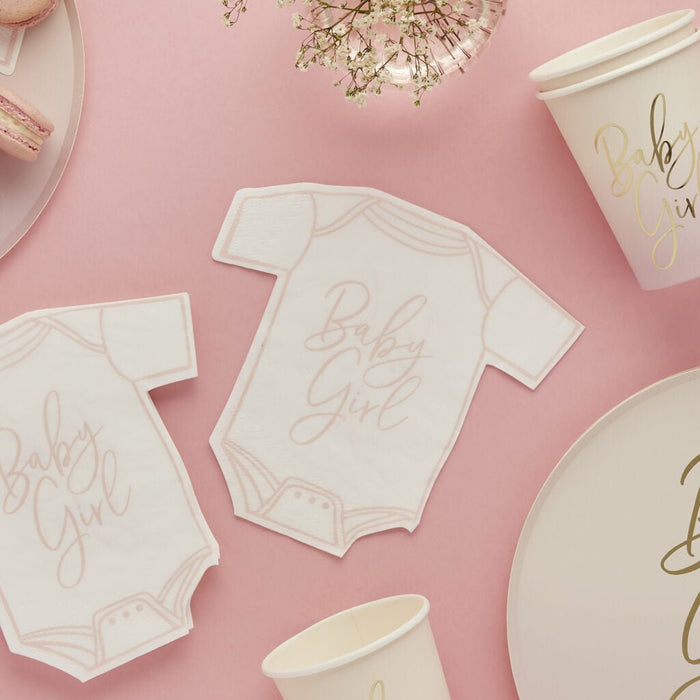 White onesie-shaped napkins with pink "Baby Girl" text arranged on a pink background with matching cups, plates, and baby's breath flowers.