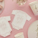White onesie-shaped napkins with pink "Baby Girl" text arranged on a pink background with matching cups, plates, and baby's breath flowers.