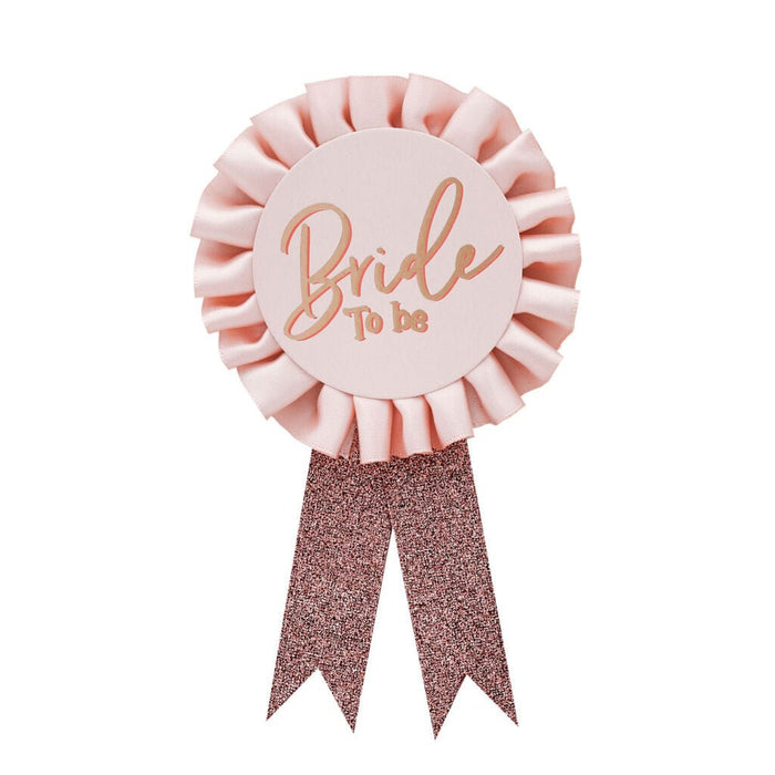 Pink rosette badge with "Bride to be" text in rose gold lettering, featuring a ruffled circular center and two glittery rose gold ribbon tails hanging below.