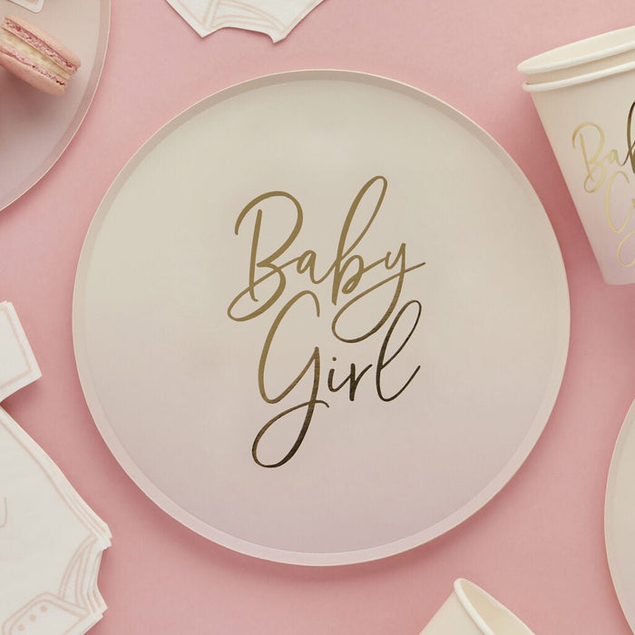 Cream-colored paper plate with gold script text reading "Baby Girl" centered on it, surrounded by matching cream and pink disposable plates and napkins on a pink background.