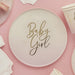 Cream-colored paper plate with gold script text reading "Baby Girl" centered on it, surrounded by matching cream and pink disposable plates and napkins on a pink background.