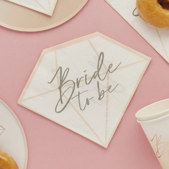 White diamond-shaped napkins with peach geometric lines and gray "Bride to be" script text, arranged on a pink background with gold-trimmed plates and pastries.