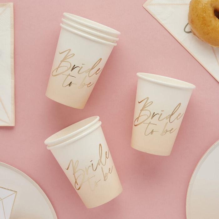 Three cream-colored paper cups with rose gold "Bride to be" script text arranged on a pink background with white napkins and a donut visible at the top.