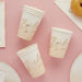Three cream-colored paper cups with rose gold "Bride to be" script text arranged on a pink background with white napkins and a donut visible at the top.