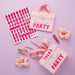 Five pink quilted pouches with hot pink text reading "Party" and "Is Las," surrounded by matching alphabet stickers, makeup brushes, nail polish, hair clips, and cream-colored hair scrunchies on a lavender background.