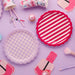 Two scalloped-edge paper plates in pink and white stripes and checkered patterns displayed on a purple background with matching party supplies including cupcakes and makeup brushes.