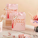 Two pink gift bags with orange heart designs and "she said yaaas" text, each tied with a coral tassel and white ribbon bow, displayed among party decorations on a wooden surface.
