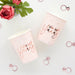 Two pink paper cups printed with "she said yaaas" text in rose gold lettering, surrounded by small decorative diamond ring props on a white background with red flowers.