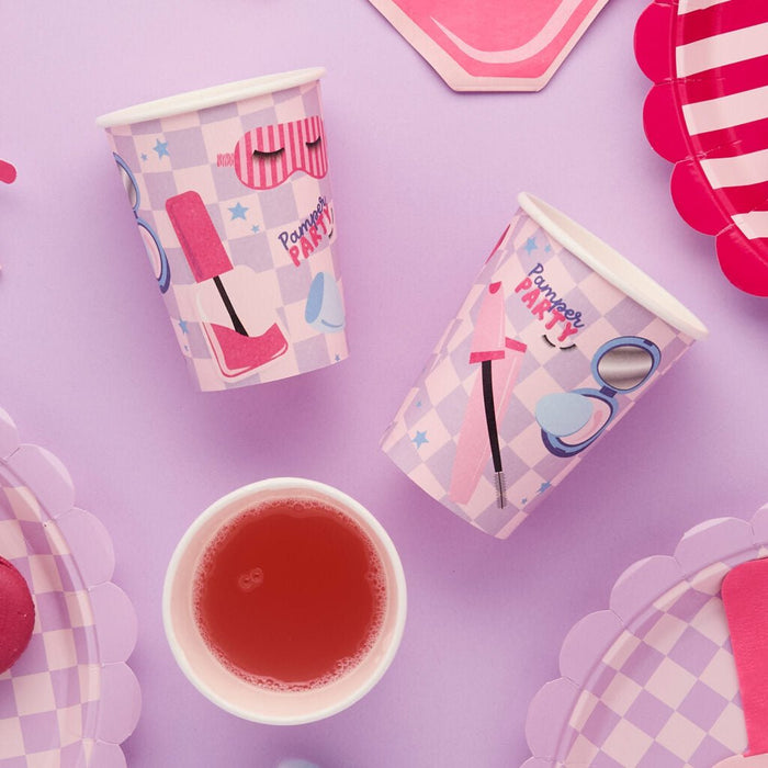 Pink and white checkered paper cups with spa-themed graphics including sleep masks and cosmetic brushes, arranged on a purple background with matching party decorations and a red beverage in the center.