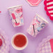Pink and white checkered paper cups with spa-themed graphics including sleep masks and cosmetic brushes, arranged on a purple background with matching party decorations and a red beverage in the center.