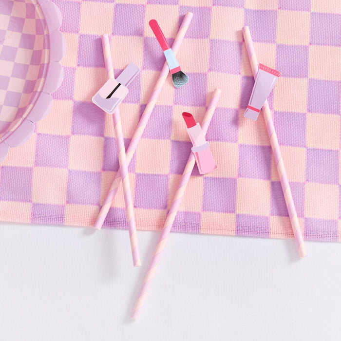 Five pink and white paper straws with cosmetic-themed designs including a lipstick, paintbrush, and nail polish bottle, arranged on a purple and pink checkered fabric background.