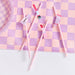 Five pink and white paper straws with cosmetic-themed designs including a lipstick, paintbrush, and nail polish bottle, arranged on a purple and pink checkered fabric background.