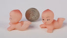 Two small peach-colored plastic baby figurines in crawling poses displayed beside a quarter coin for size reference.
