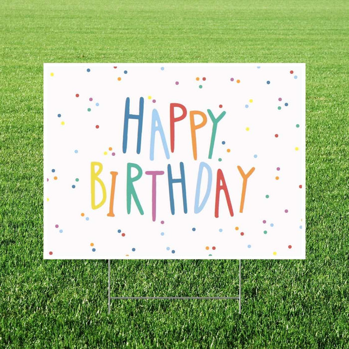 Colorful "Happy Birthday" yard sign with multicolored letters and confetti dots on white background, displayed on grass with metal stakes.