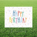 Colorful "Happy Birthday" yard sign with multicolored letters and confetti dots on white background, displayed on grass with metal stakes.