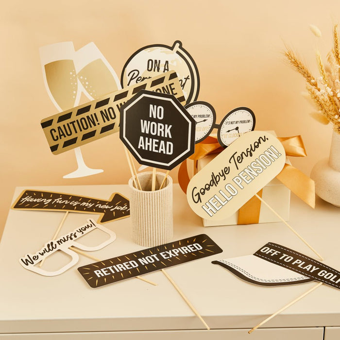 Retirement party photo booth props set with ten black and cream decorative signs on sticks, including champagne glasses, caution tape, clocks, and humorous messages like "No Work Ahead" and "Retired Not Expired" arranged on a beige surface with wheat decorations.