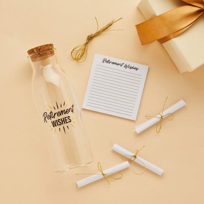 Glass bottle with cork stopper labeled "Retirement Wishes" in black text with gold sun rays, surrounded by lined notepad, rolled message scrolls tied with gold string, and gold decorative bows on cream background.