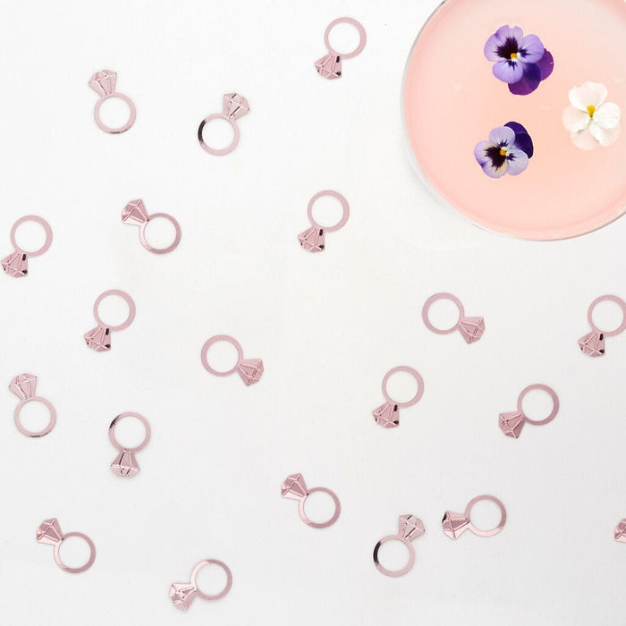 Multiple rose gold diamond-shaped ring charms scattered on white background with a pink circular plate containing purple and white pansies in the upper right corner.