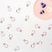 Multiple rose gold diamond-shaped ring charms scattered on white background with a pink circular plate containing purple and white pansies in the upper right corner.