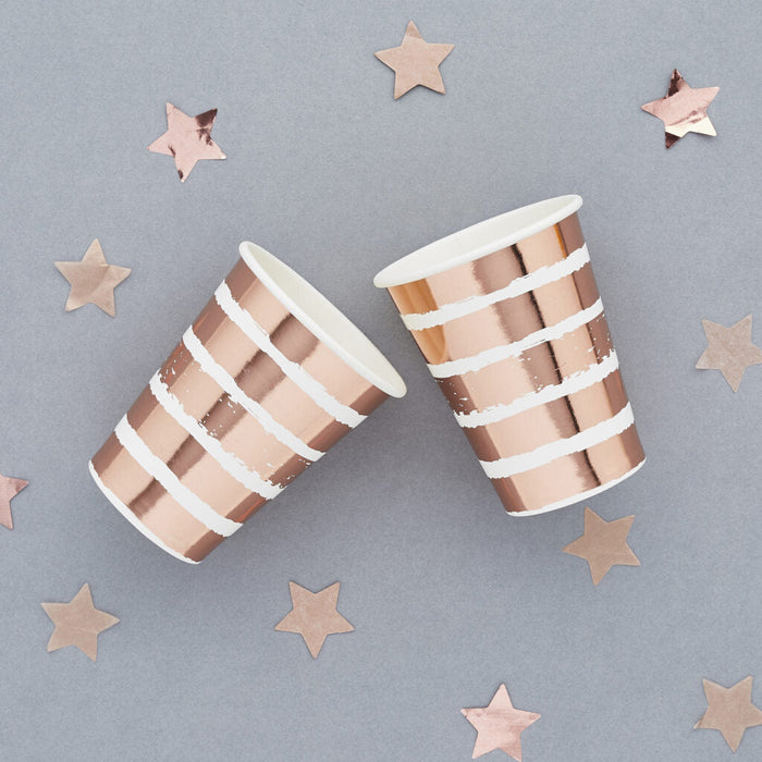 Two rose gold and white striped paper cups surrounded by rose gold star decorations on a gray background.
