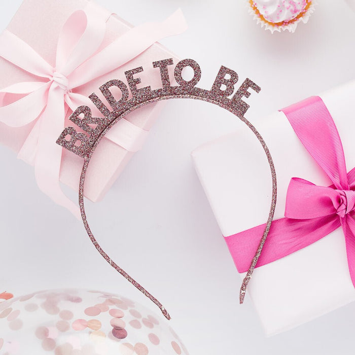 Rose gold glitter "Bride to Be" headband with pink ribbon bows displayed on white background surrounded by pink decorative elements.