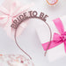 Rose gold glitter "Bride to Be" headband with pink ribbon bows displayed on white background surrounded by pink decorative elements.