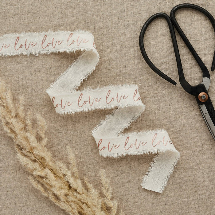 Three white cotton ribbons with rose gold "love" text and frayed edges displayed on a beige linen background with dried pampas grass and black scissors.