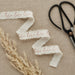 Three white cotton ribbons with rose gold "love" text and frayed edges displayed on a beige linen background with dried pampas grass and black scissors.