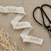Three white cotton ribbons with frayed edges and rose gold "Mr & Mrs" text printed on them, arranged on a beige linen background next to black scissors with a copper detail.
