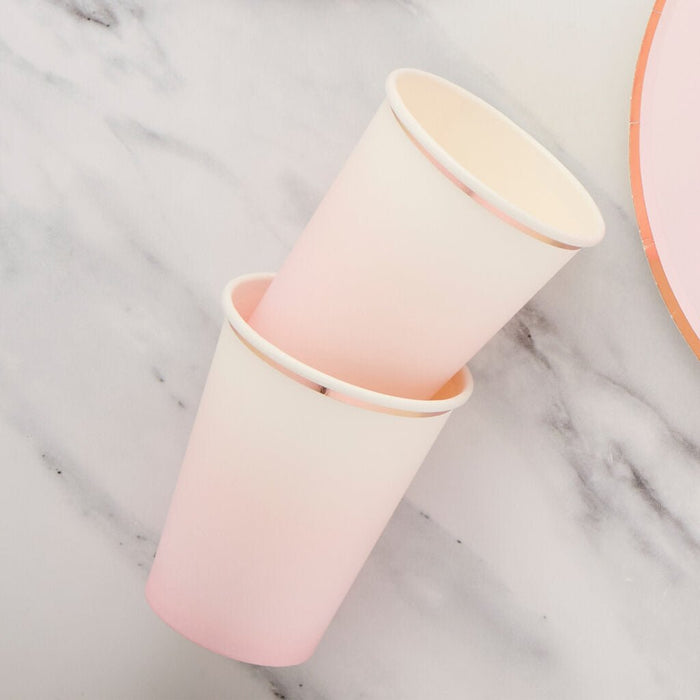 Two nested pink ombre paper cups with cream-colored rims displayed at an angle on a marble surface with an orange ribbon visible on the right.
