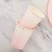 Two nested pink ombre paper cups with cream-colored rims displayed at an angle on a marble surface with an orange ribbon visible on the right.