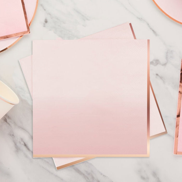 Stack of light pink paper napkins with rose gold ombre gradient and metallic gold trim, arranged on a marble surface with copper ribbon accents.