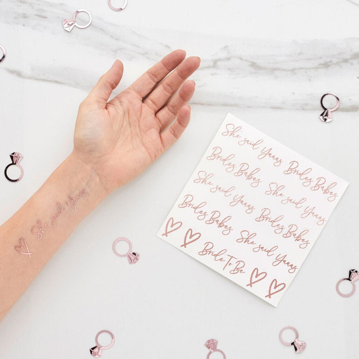 Hand displaying rose gold temporary tattoos with "She Said Yass," "Bride," and "Baby" text and hearts, alongside a sheet of matching tattoo designs and decorative ring charms on marble background.