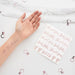Hand displaying rose gold temporary tattoos with "She Said Yass," "Bride," and "Baby" text and hearts, alongside a sheet of matching tattoo designs and decorative ring charms on marble background.