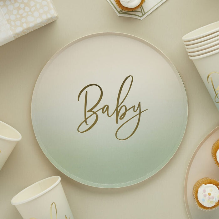 Large round paper plate with "Baby" written in gold script on a sage green ombre background, surrounded by coordinating white cups, napkins, and cupcakes in a flat lay arrangement.