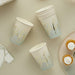 Eight cream and sage ombre paper cups with gold "Baby" script text, displayed with matching napkins and frosted cupcakes on a beige surface.