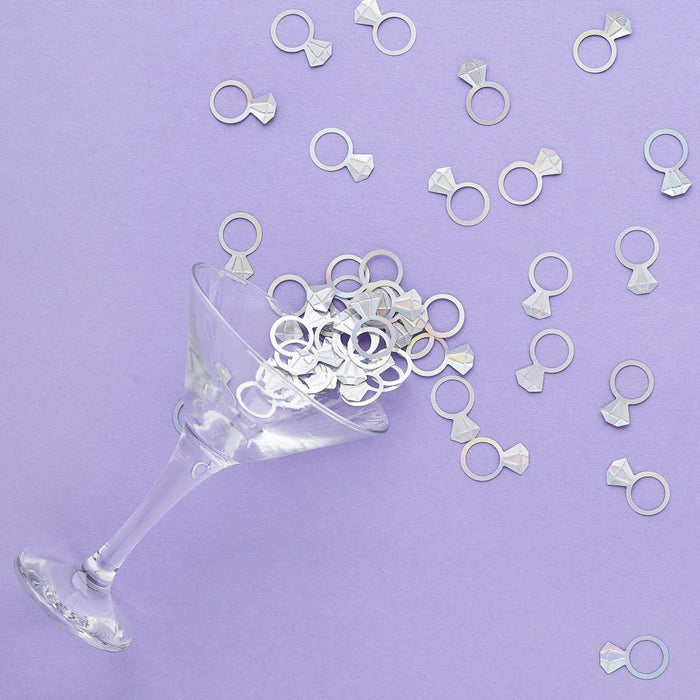 Silver ring-shaped confetti pieces with bow tie details scattered across a purple background and overflowing from a clear martini glass.