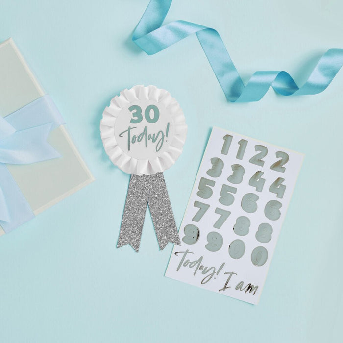 White rosette badge with silver text reading "30 Today!" and matching silver glitter ribbon tails, displayed on a light blue background with a scratch-off card and blue ribbon accents.