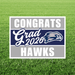 Yard sign with gray and navy horizontal stripes displaying "CONGRATS Grad 2026 HAWKS" text and a hawk mascot logo on green grass background.