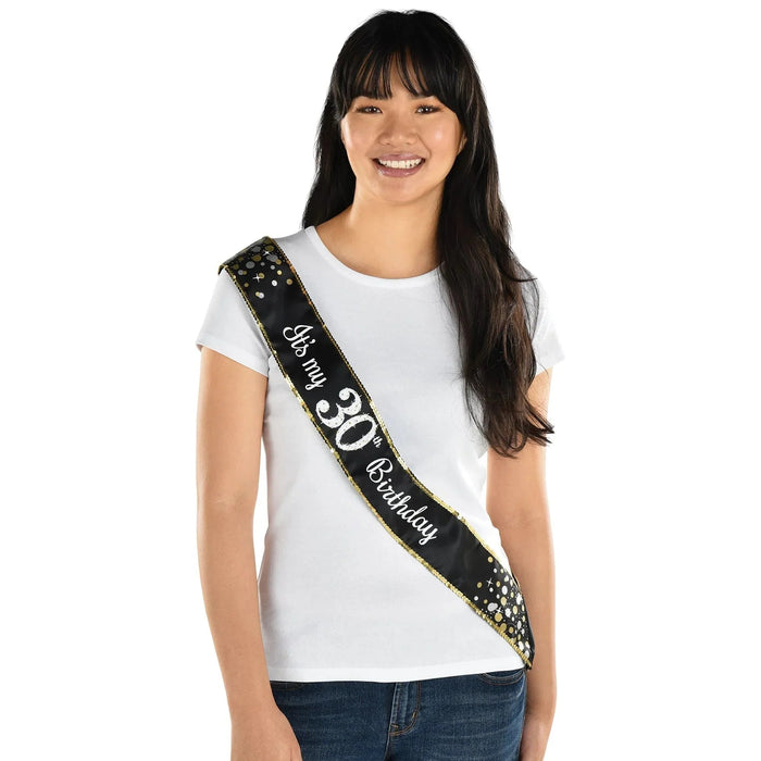 Black fabric sash with gold sequin trim and white text reading "30 with Birthday" worn diagonally across a white t-shirt on a smiling young woman.