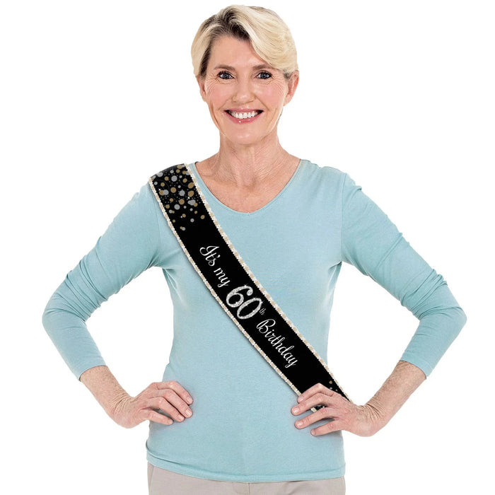 Black fabric sash with gold studs and "It's my 60th Birthday" text in white, displayed on a woman wearing a light blue long-sleeved shirt.