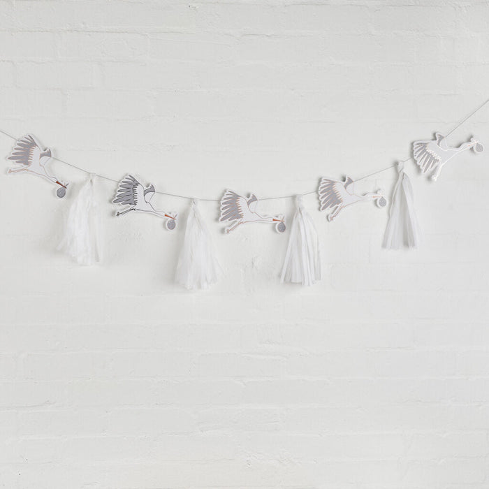 White stork decorations with gray wing details and orange beaks are suspended on a string with white tissue paper tassels hanging below, creating a delicate baby shower garland against a white brick wall.