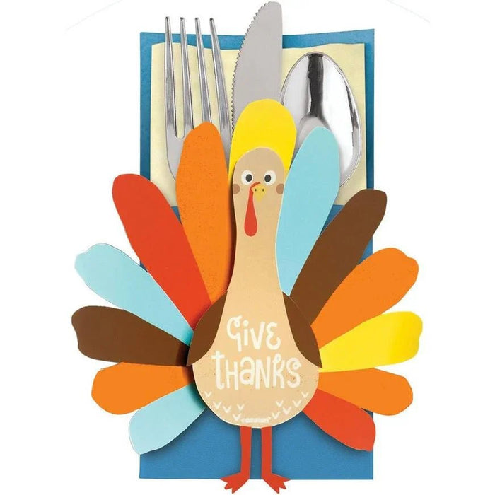 Colorful paper turkey-shaped utensil holder with "Give Thanks" text, featuring a cream body, yellow head, red beak, and multicolored feathers in orange, red, yellow, blue, and brown, holding a fork, knife, and spoon against a blue background.